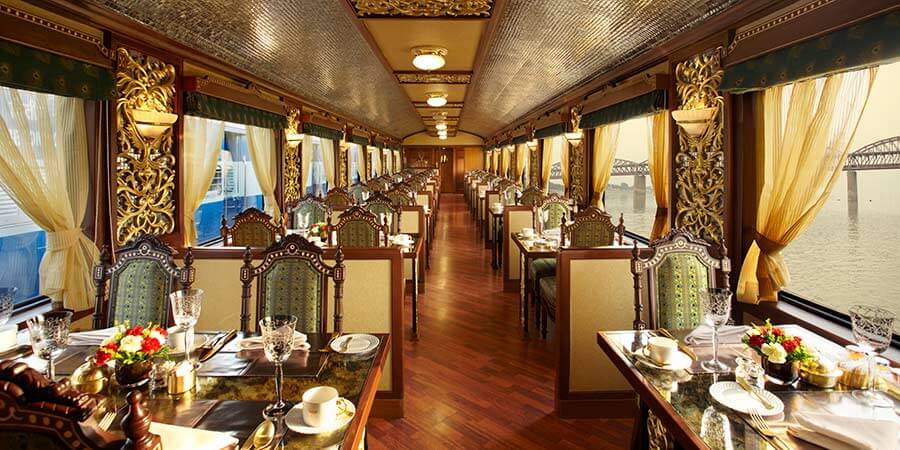 View down the dining cart of the Maharajas’ Express adorned with ornate wooden panelling, golden accents, and intricate lighting fixtures, creating a regal and cozy atmosphere. Antique-style chairs at each table add to the vintage charm of the train's design.