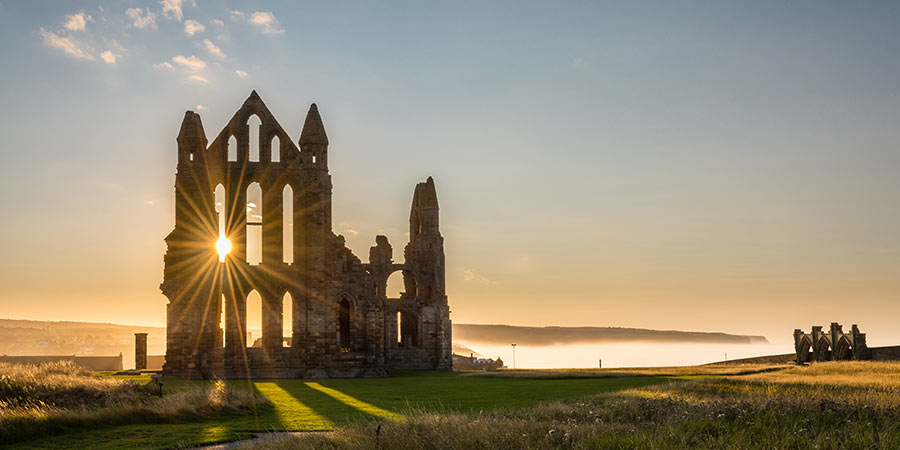 Sun shines through an old Abbey at sunset, behind is mist covering the sea.