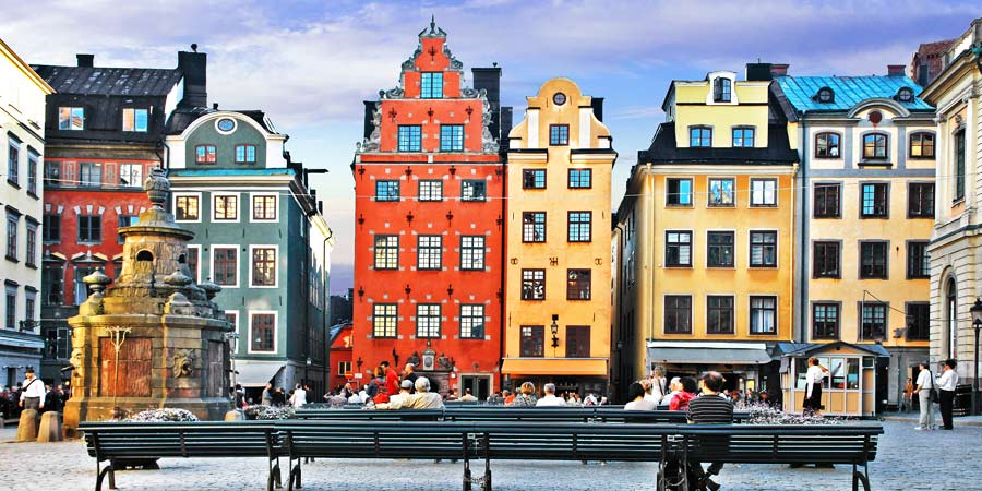 Tourists enjoy the traditional architecture of Stockholm’s old town buildings. The houses are pale yellow and bright red with many windows and ornate roof designs.