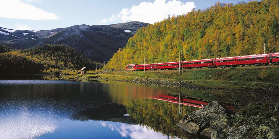 A bright red train passes by a lake with autumnal trees behind.