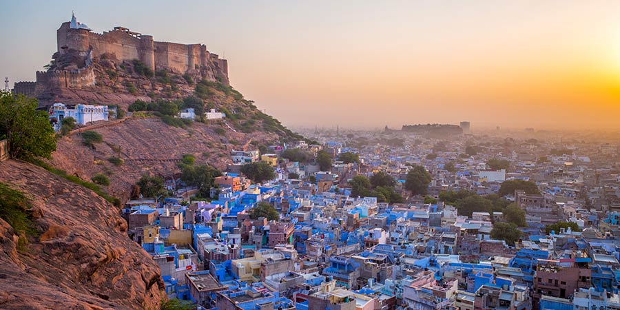 A panoramic view of Jodhpur, located within Rajasthan, known as the 'Blue City,' with its iconic blue-painted houses scattered across the landscape. The majestic Mehrangarh Fort stands tall atop the hill, overlooking the city below. The vibrant streets are filled with traditional Rajasthani architecture and narrow alleys at sunset